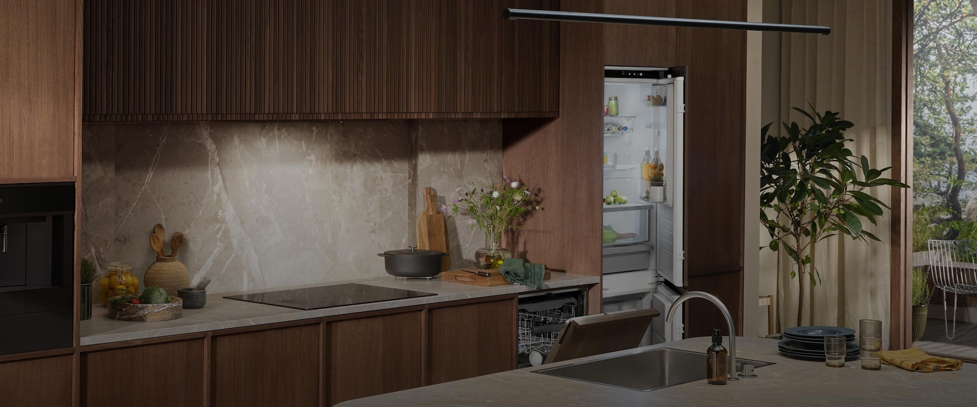 Integreret kølefryseskab og opvaskemaskine i et stilrent køkken med mørke træpaneler og bordplade i marmor.